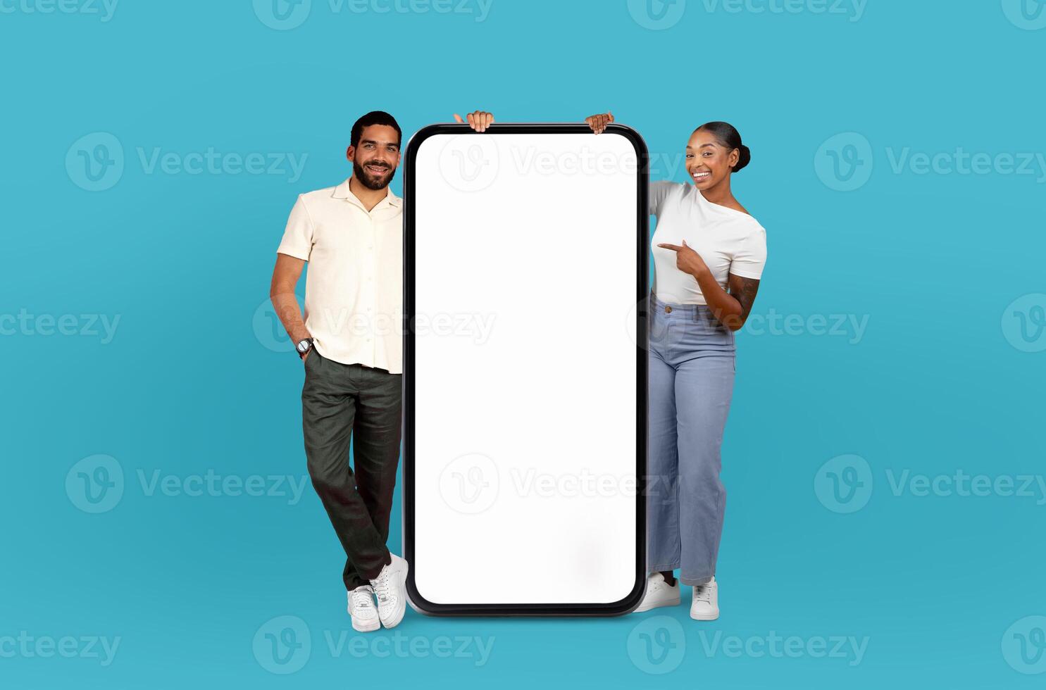 A cheerful hispanic man and black woman are standing against a turquoise backdrop, presenting a giant, blank, vertical smartphone screen. photo