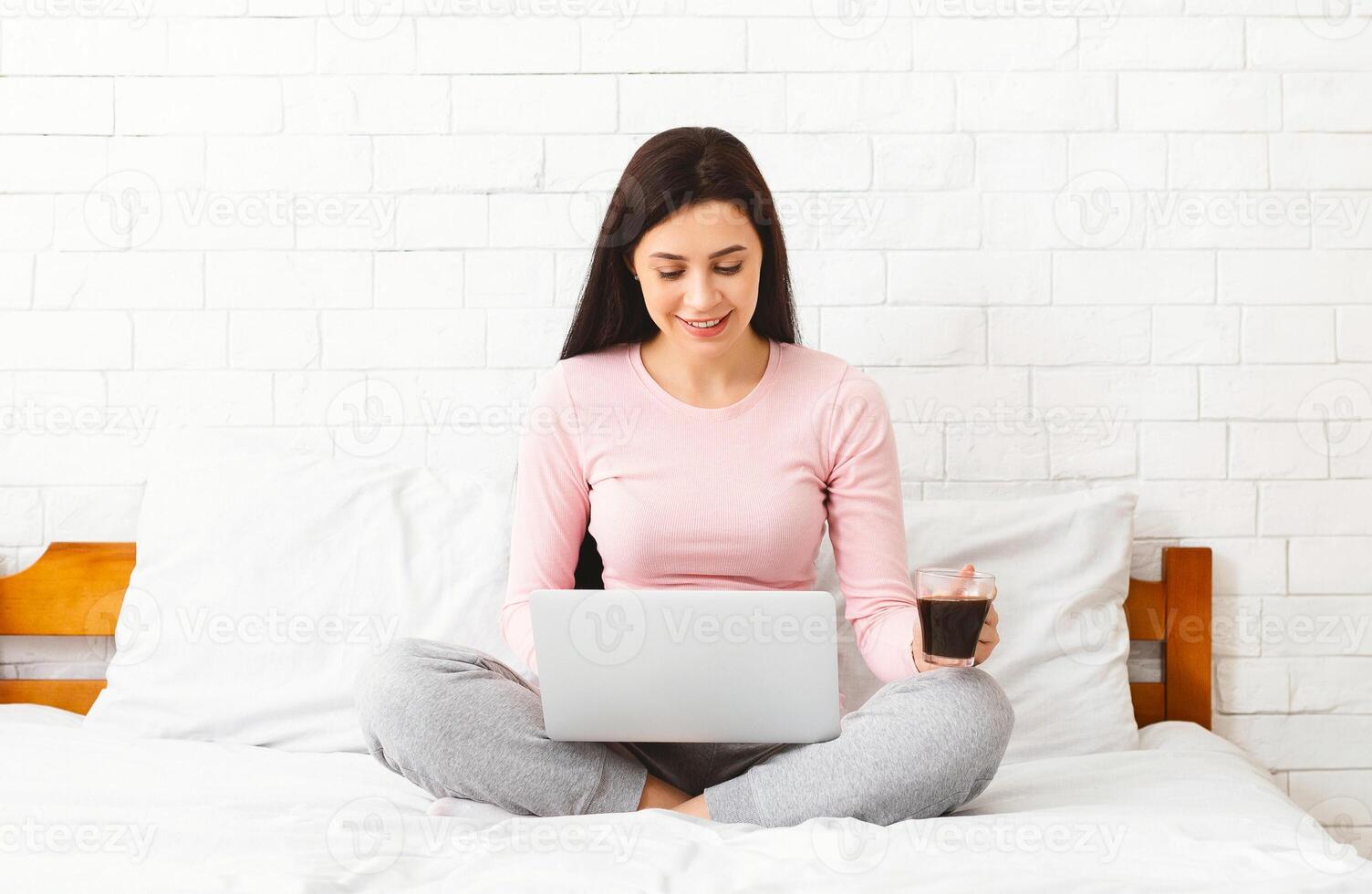 A woman is sitting on a bed, working on a laptop while holding a cup of coffee. She seems focused and engaged in her work, with the morning light filtering through the window. photo