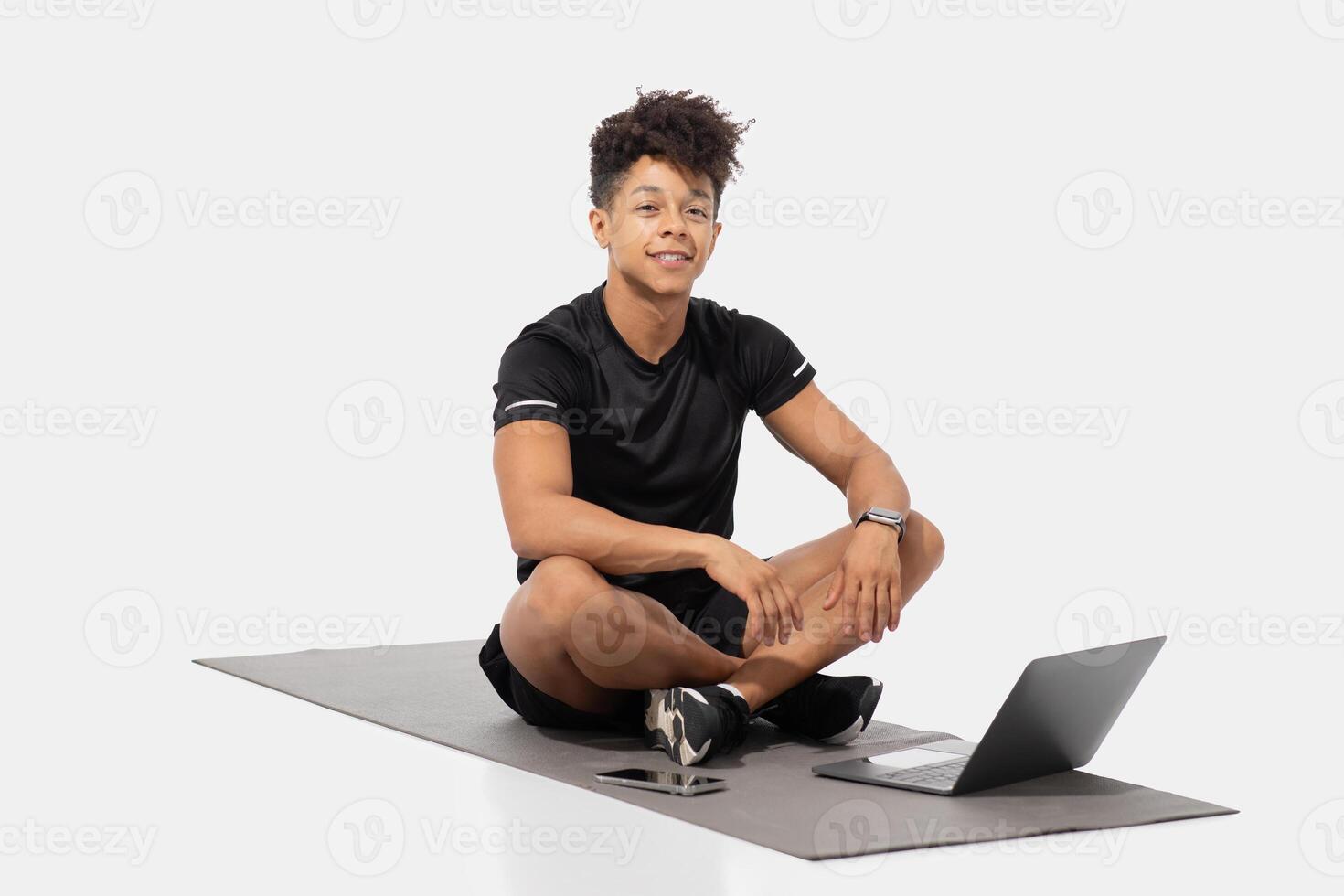 A young man sits cross-legged on a mat while participating in an online fitness class. He is focused on the screen of his laptop, which displays workout instructions photo