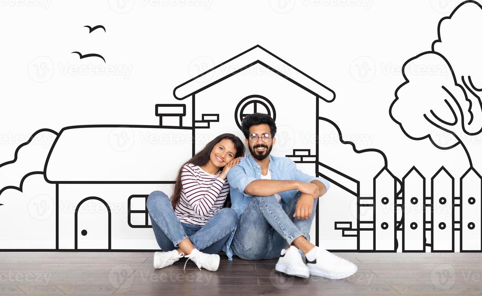 Cheerful young indian couple sitting by white wall with a house outline, dreaming of a home. Real estate, mortgage concept photo