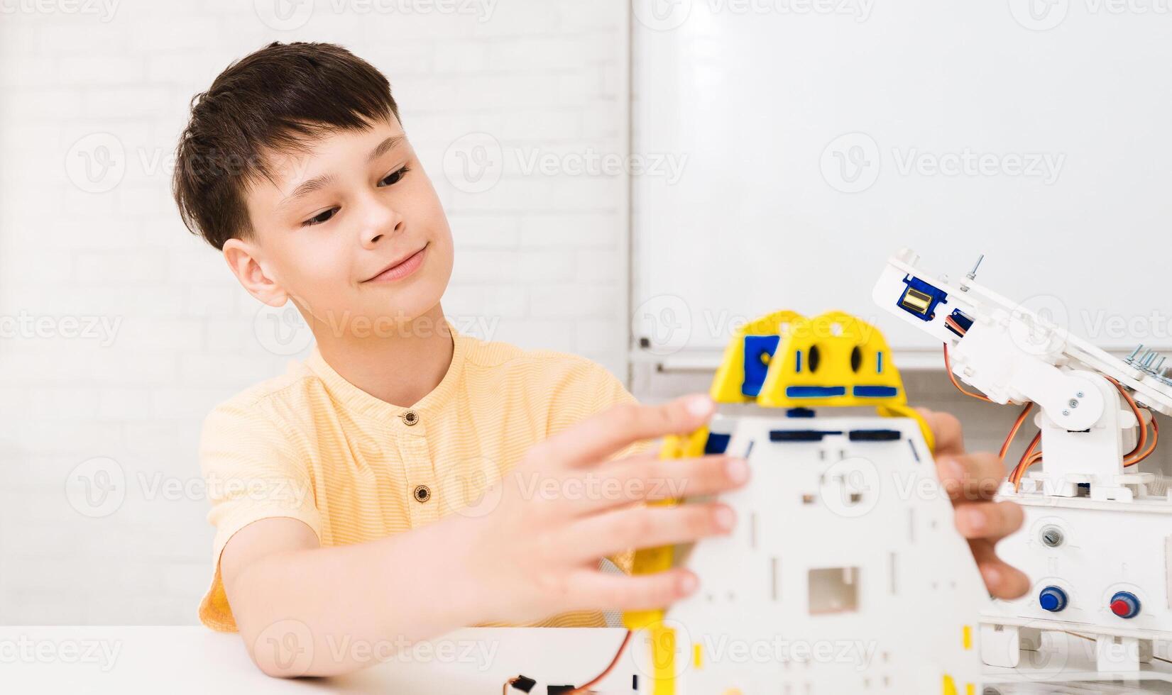A young boy with short brown hair sits at a table, building a robot. He is wearing a yellow shirt and has a concentrated look on his face as he works on the robot photo