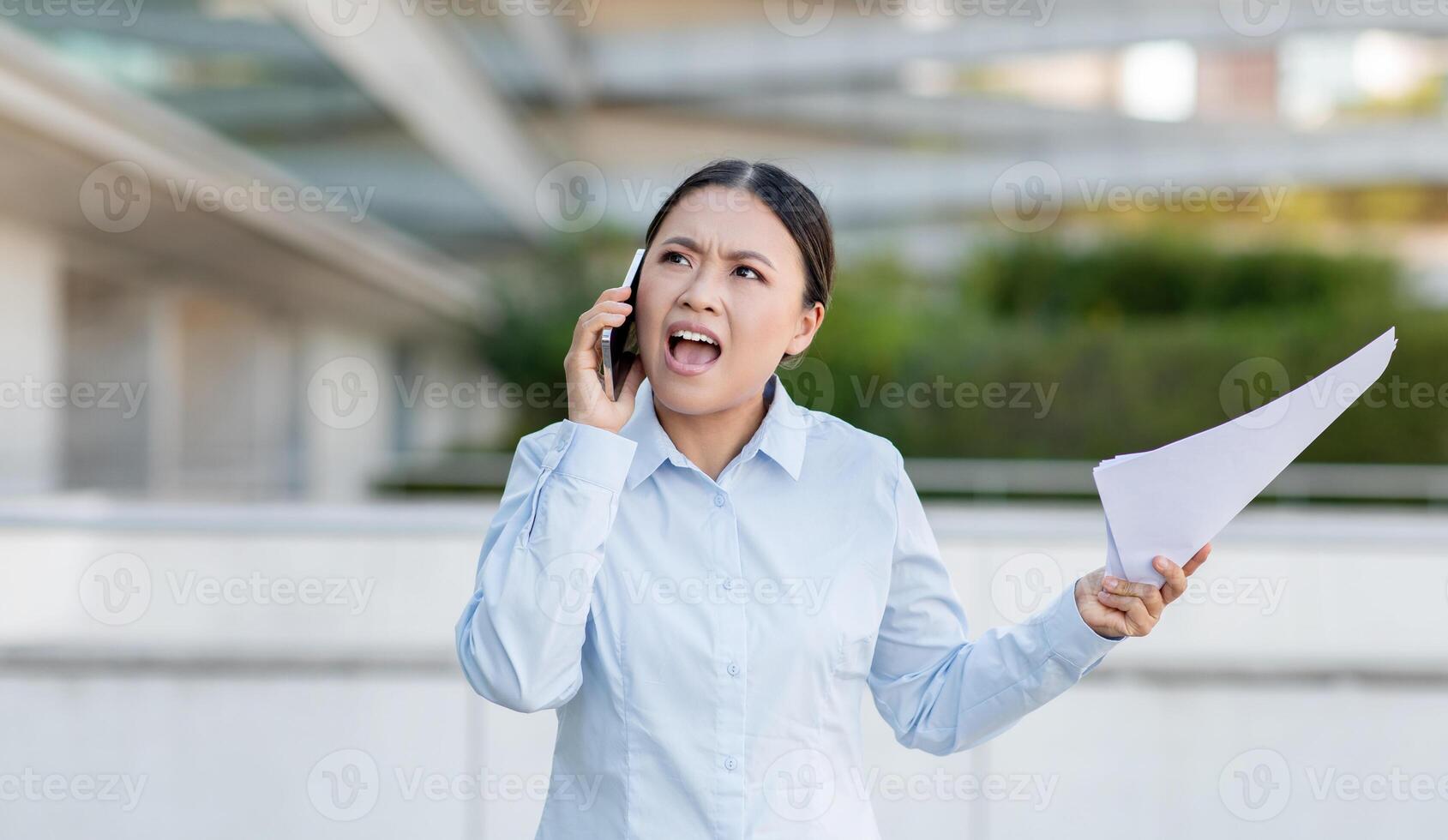 Asian woman dressed in a light blue shirt appears distressed as she talks on her phone. She is outdoors, gesturing with her left hand while clutching several documents in her right. photo