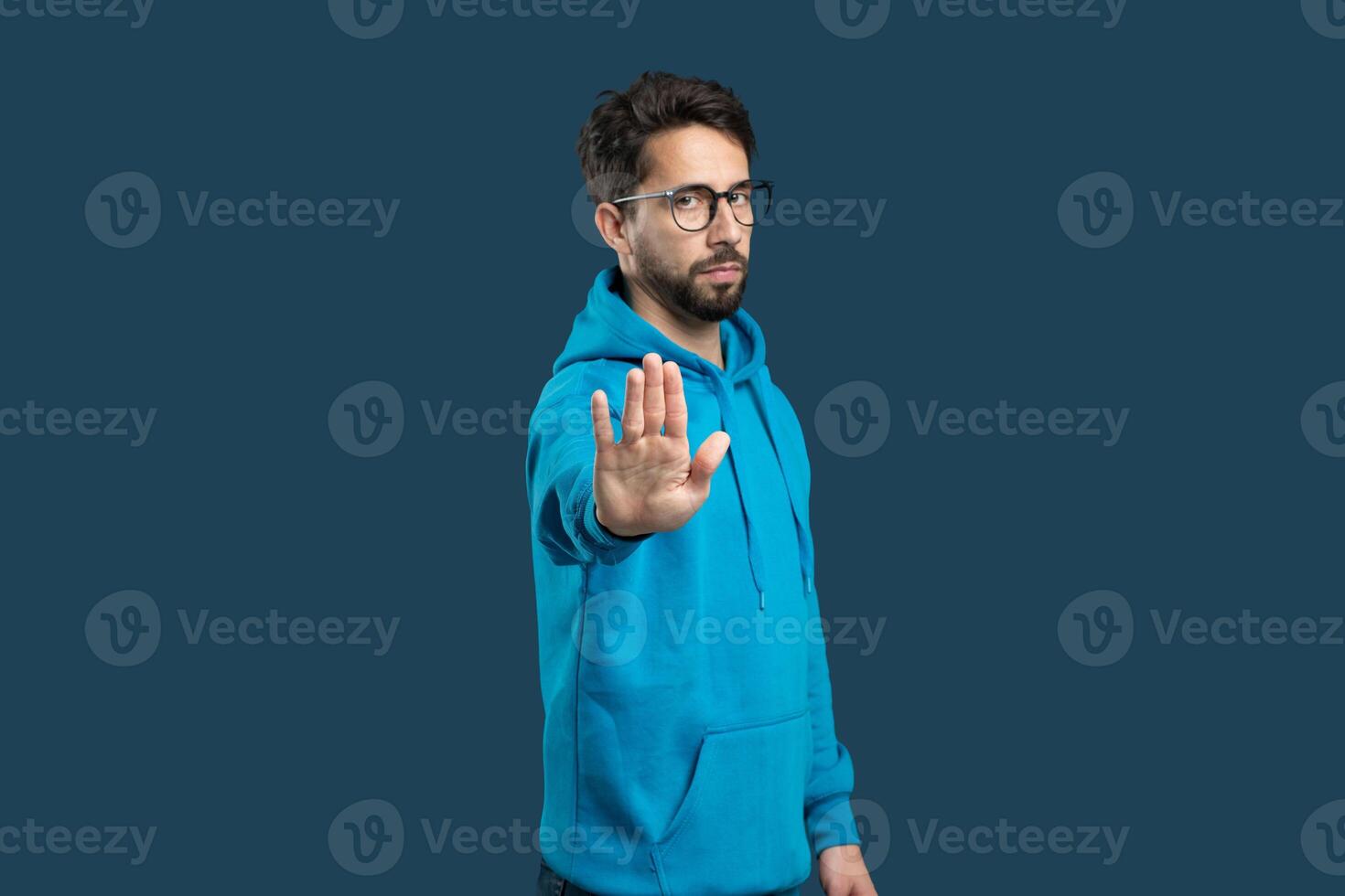 A young man stands confidently, wearing a bright blue hoodie and glasses. He holds his hand up in a gesture that conveys a message of pause or stop. The solid background photo