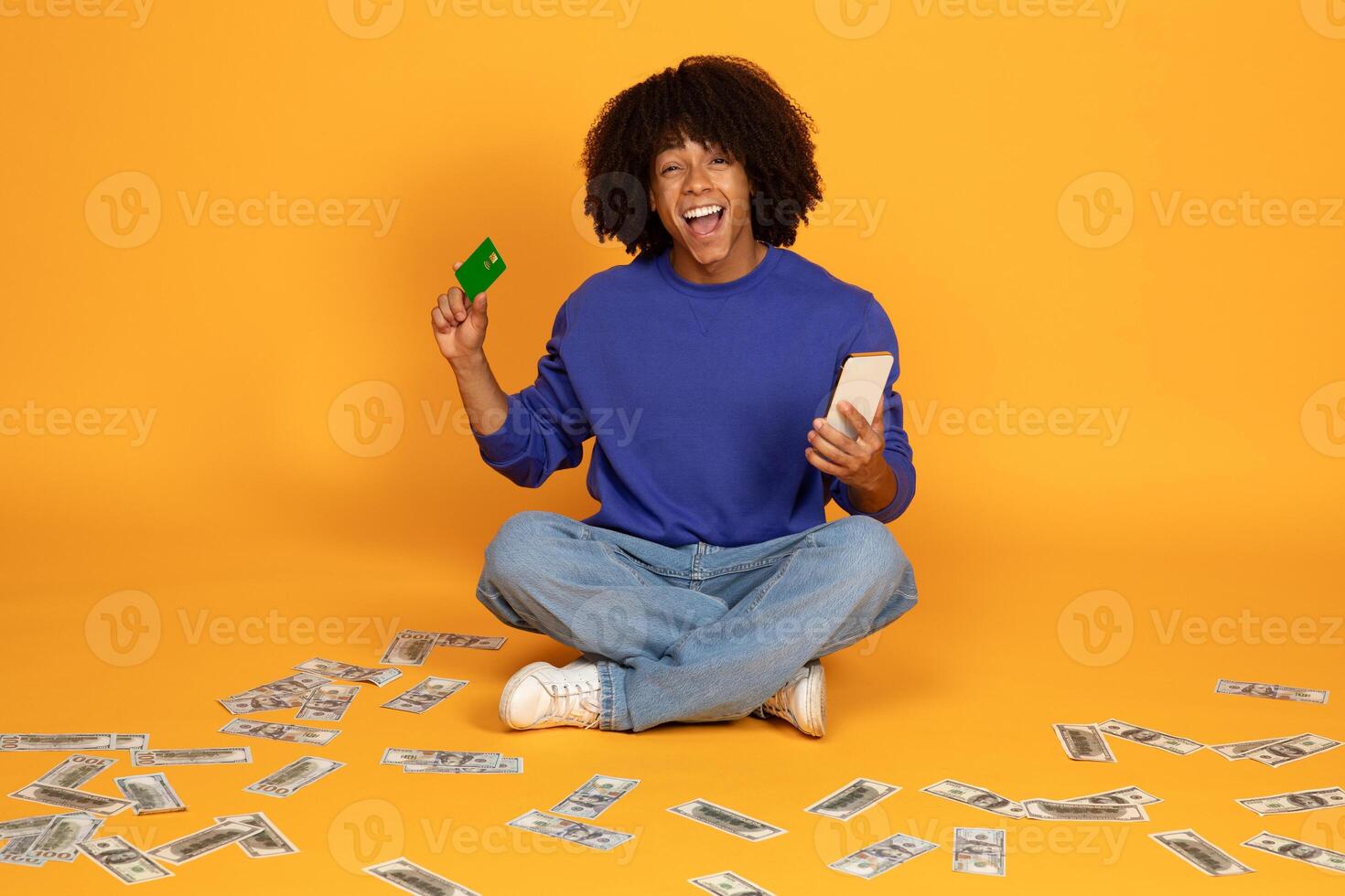 A young man with an curly sits on the floor with his legs crossed, holding a credit card in one hand and a smartphone in the other. The floor is covered in scattered cash. photo