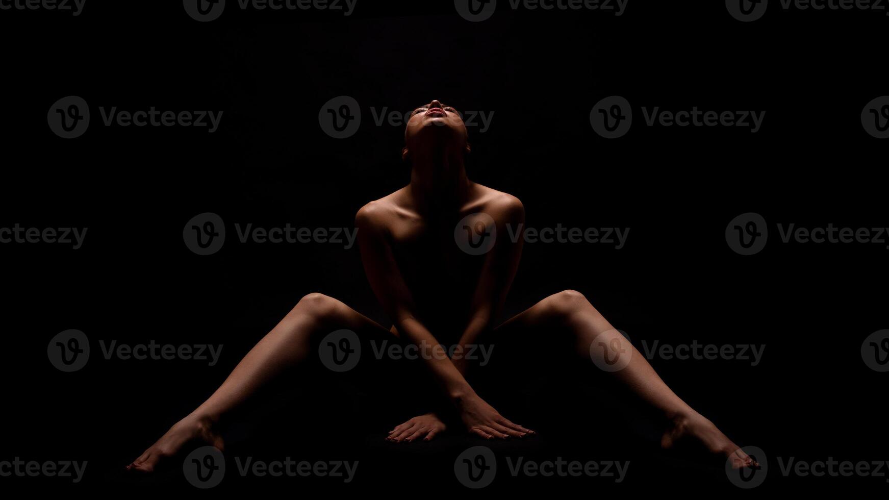 A woman sits in a dark room, her head tilted back and her legs spread out. She is bare-chested, and the light from above illuminates her face and body. photo