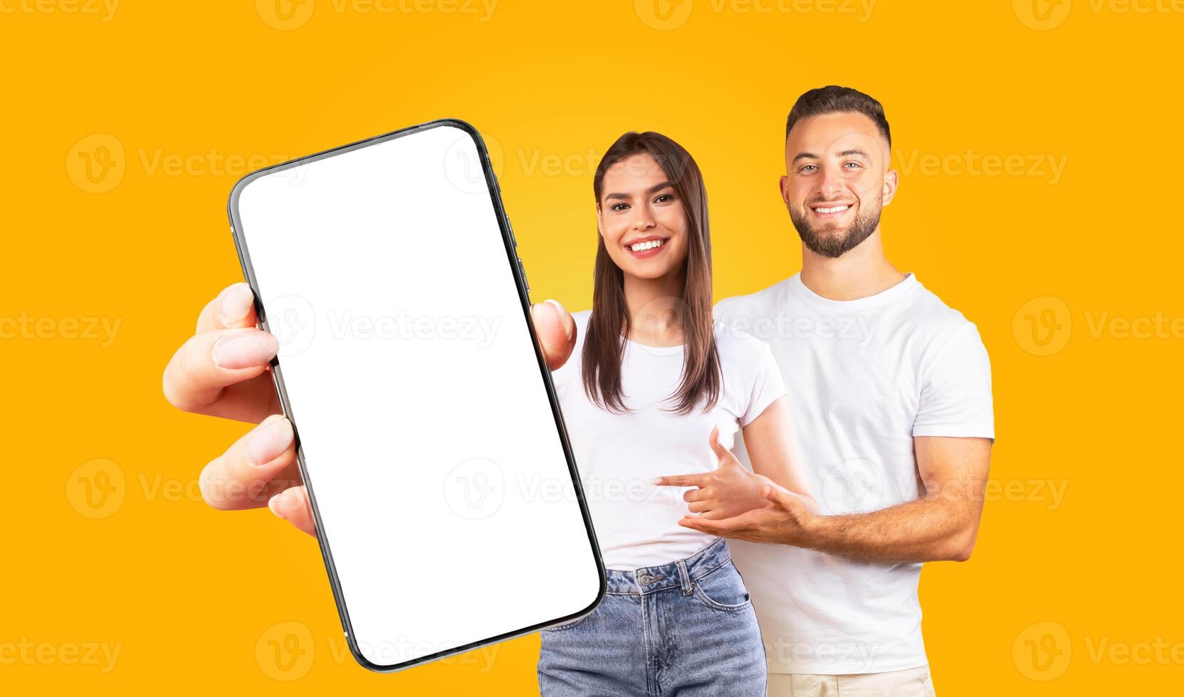 A young couple is holding up a smartphone with a blank screen. The man and woman are smiling and looking at the camera. They are both wearing casual clothing photo