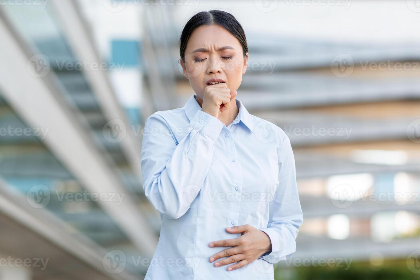 A young Asian woman stands outside a contemporary building, appearing unwell. She clutches her stomach with one hand while coughing, expressing discomfort photo