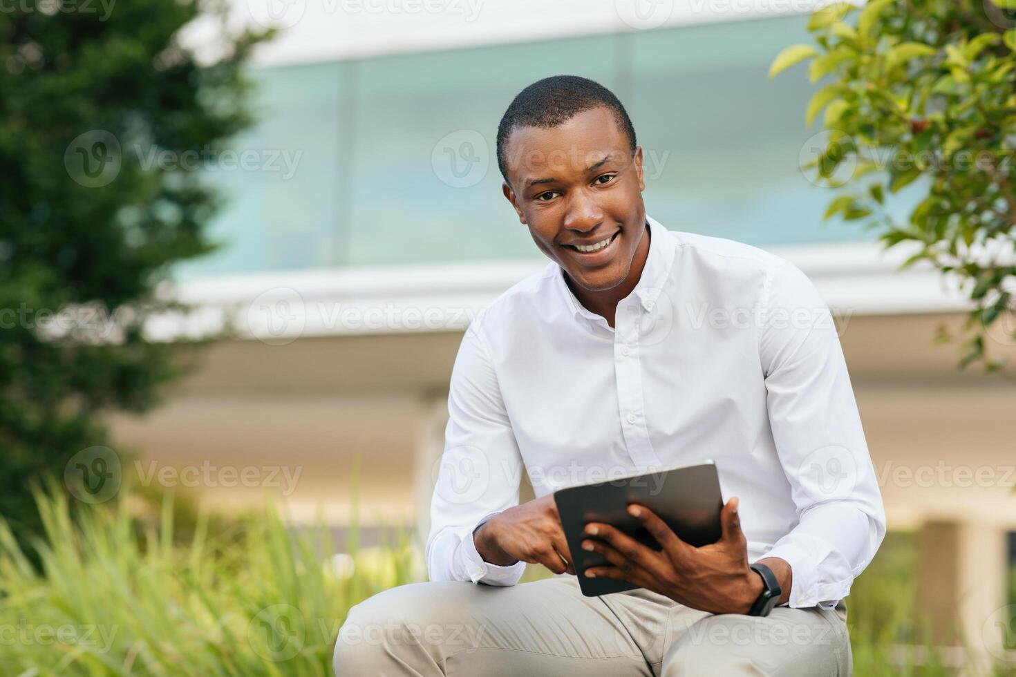 A young African American man sits outdoors on a bench, wearing a white button-down shirt and beige pants, and uses a tablet. He is smiling and looking directly at the camera photo