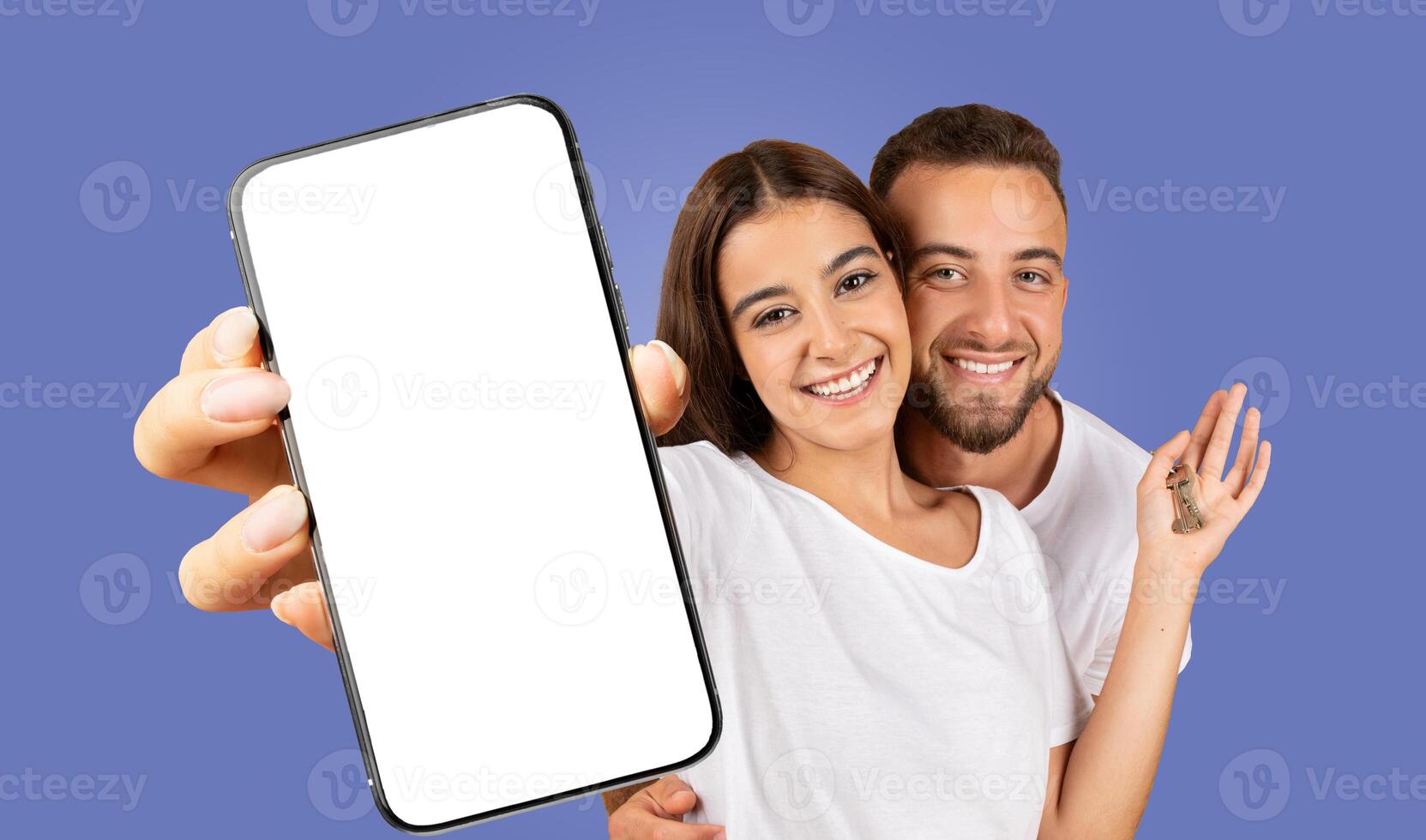 A young couple stands in front of a solid blue background, smiling brightly at the camera. The woman is holding a smartphone with a blank screen photo