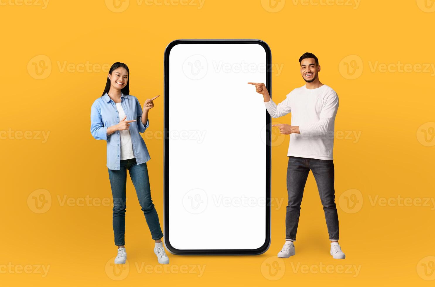 The scene features a young multiethnic couple, both grinning, positioned next to a life-sized smartphone mockup on a seamless yellow background. photo