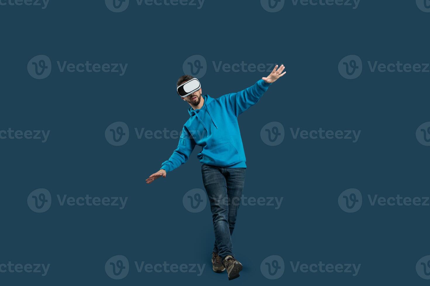 A young man engages in a balance exercise while wearing a virtual reality headset. He stands on one leg, arms outstretched for stability, in a modern indoor setting with a solid color backdrop photo