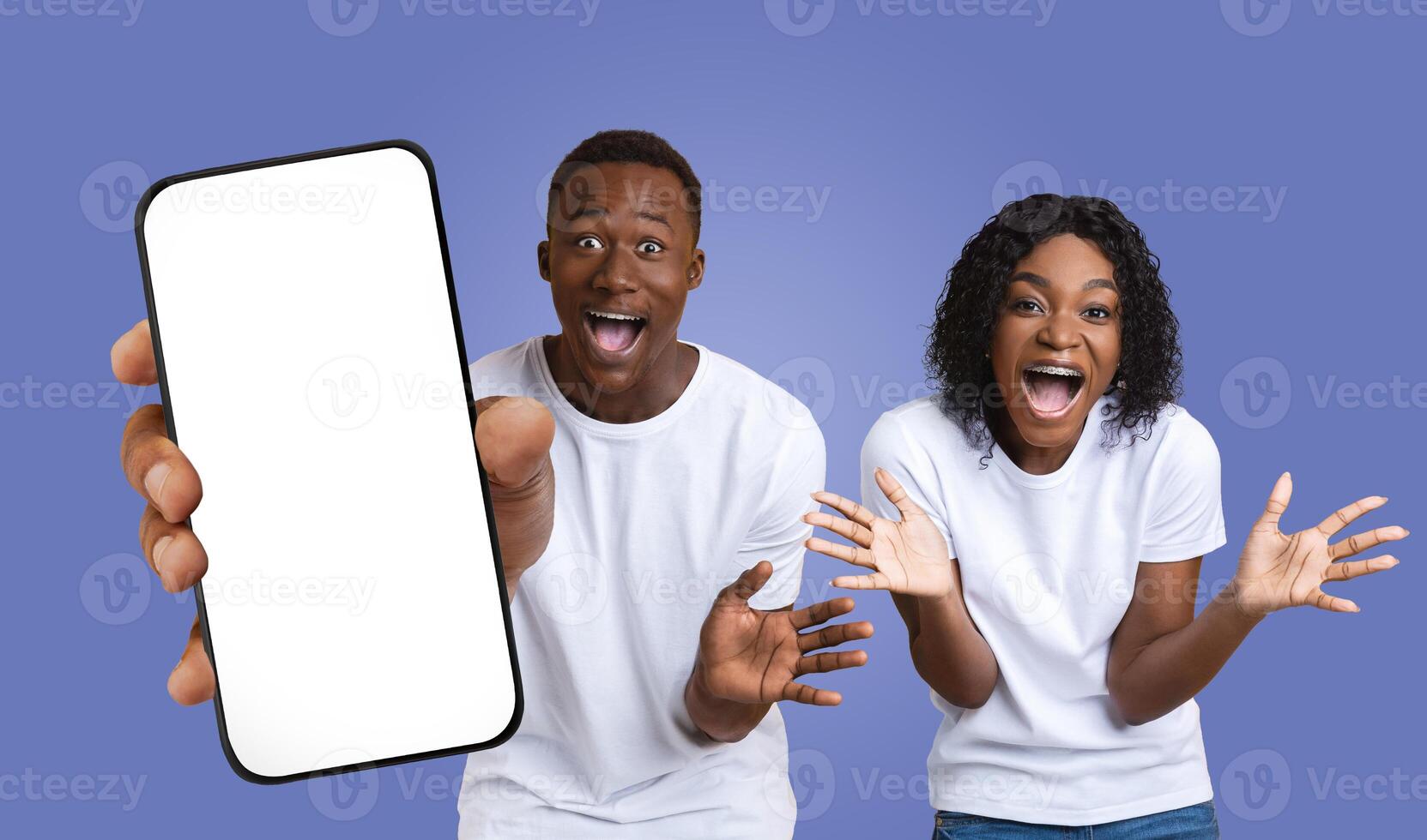 A young black couple, dressed in casual white shirts, express their excitement while holding a smartphone with a blank screen. The background is a solid purple color. photo