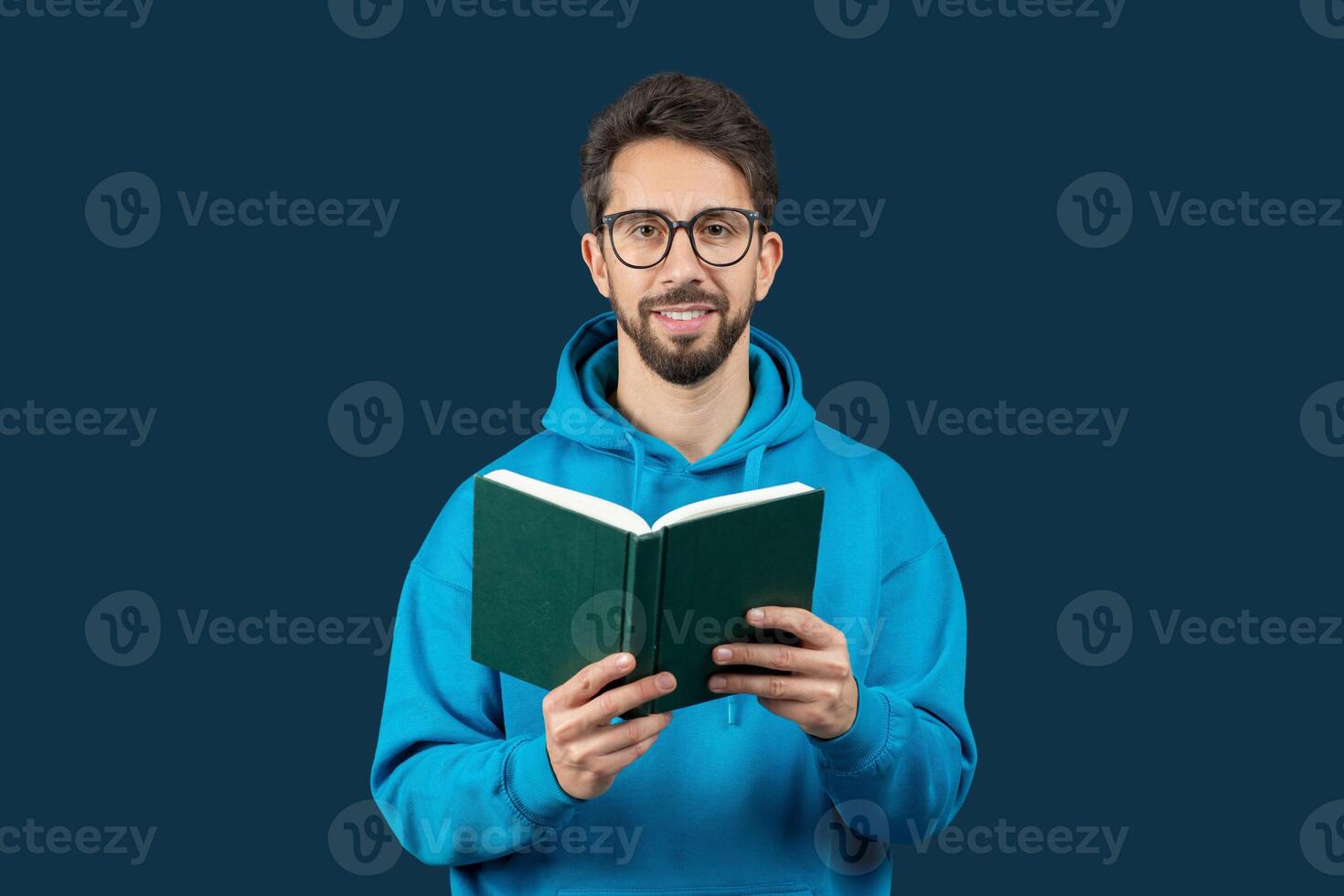 A man wearing a blue hoodie is engrossed in reading a book. He sits indoors, focused on the pages, with a calm demeanor as he flips through the book. photo