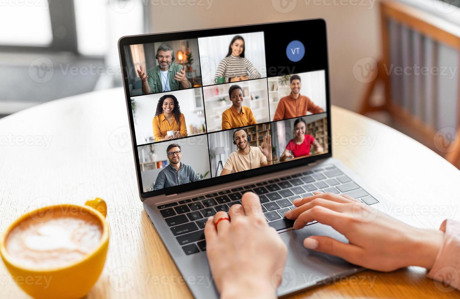 Lady sits at a table in a coffee shop, using a laptop to participate in a conference with a group of people. The screen shows six tiles of the participants photo