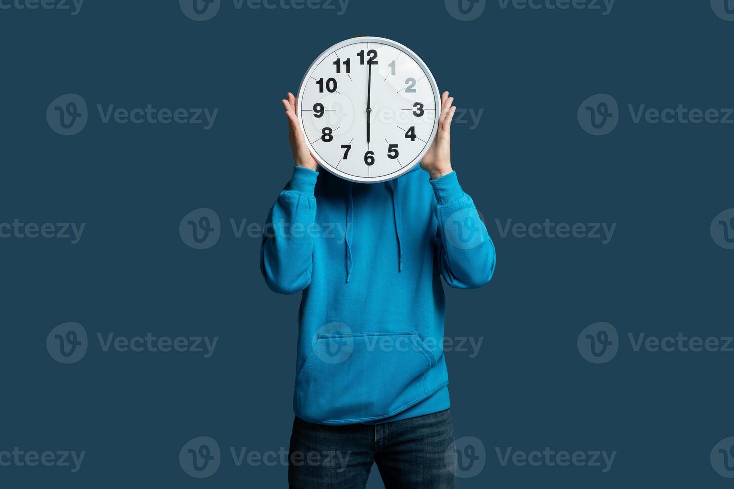 A young man stands against a blue wall, holding a large wall clock in front of their face. He is dressed in a blue hoodie and jeans, creating a playful and humorous visual representation of time photo