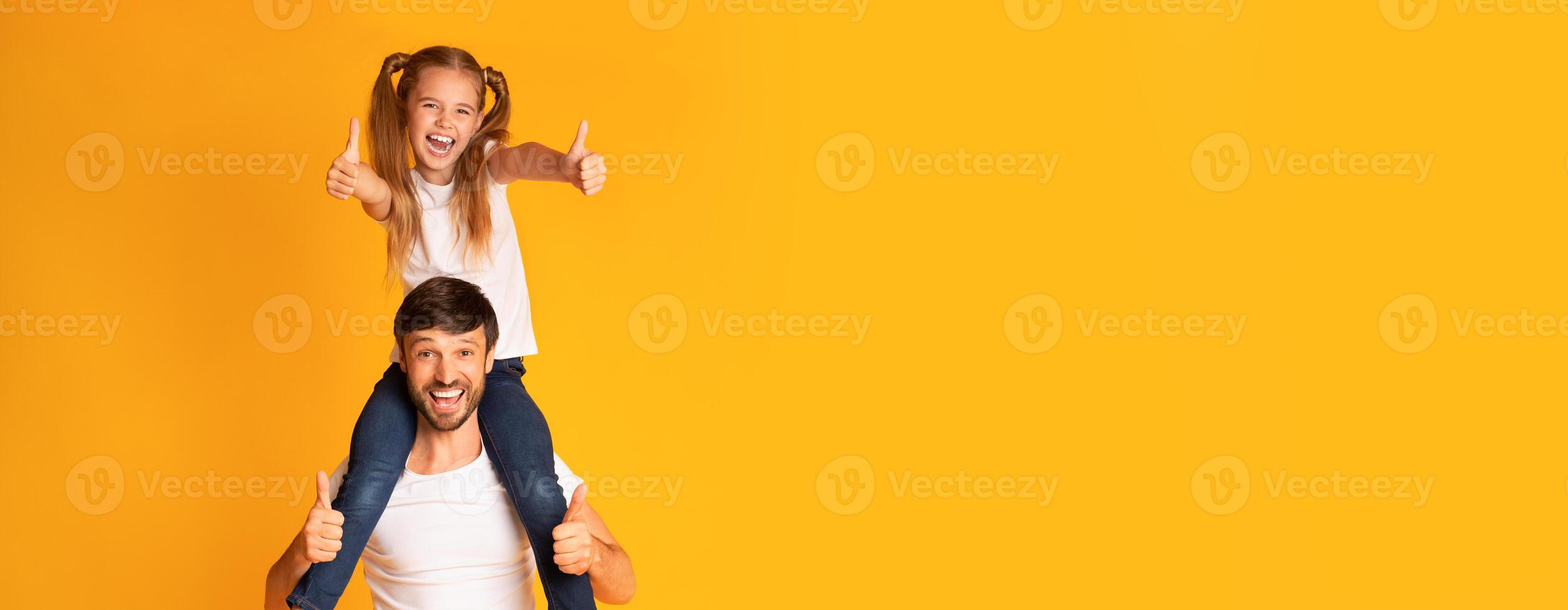 A joyful father lifts his smiling daughter onto his shoulders, both giving thumbs up with cheerful expressions. The bright yellow backdrop enhances the playful atmosphere, copy space photo