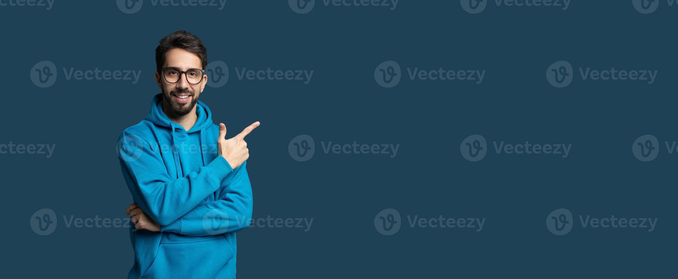 A young man wearing a blue hoodie stands confidently with his arms crossed and a smile on his face. He points at copy space, engaging viewers attentively. The background is a simple, solid color photo