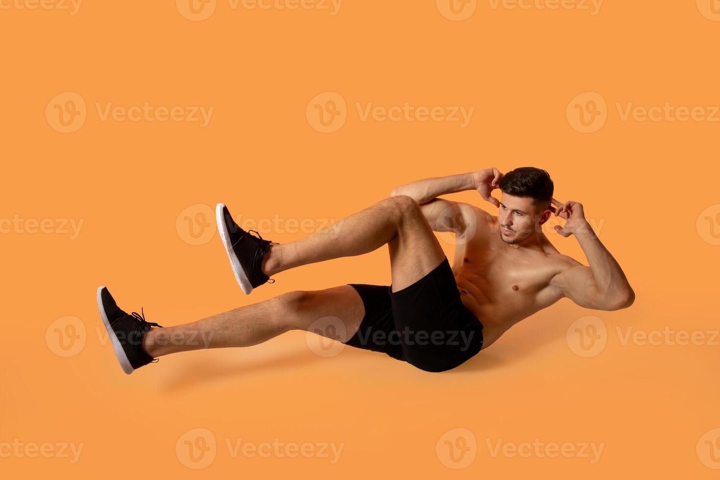 A fit man in black shorts and sneakers is performing a bicycle crunch exercise on an orange studio background. He is focused and demonstrating proper form. photo