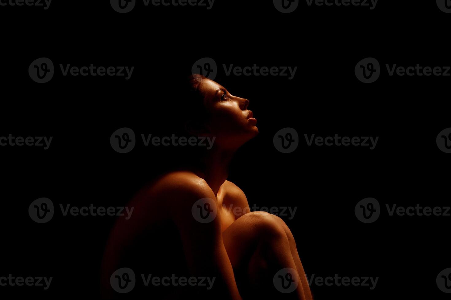 The image shows a woman sitting in a dark room. She is illuminated by a warm, golden light that highlights her face, shoulders, and upper body. photo