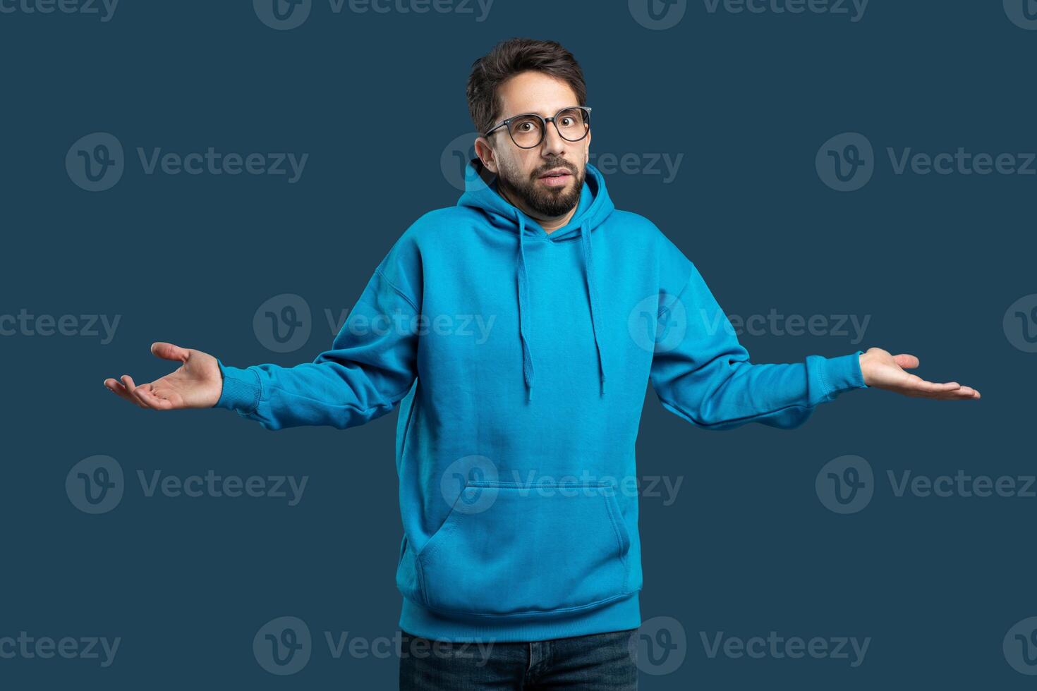 Confused man wearing a blue hoodie is standing with his hands outstretched. His body language suggests he may be gesturing or expressing something. The background is simple photo