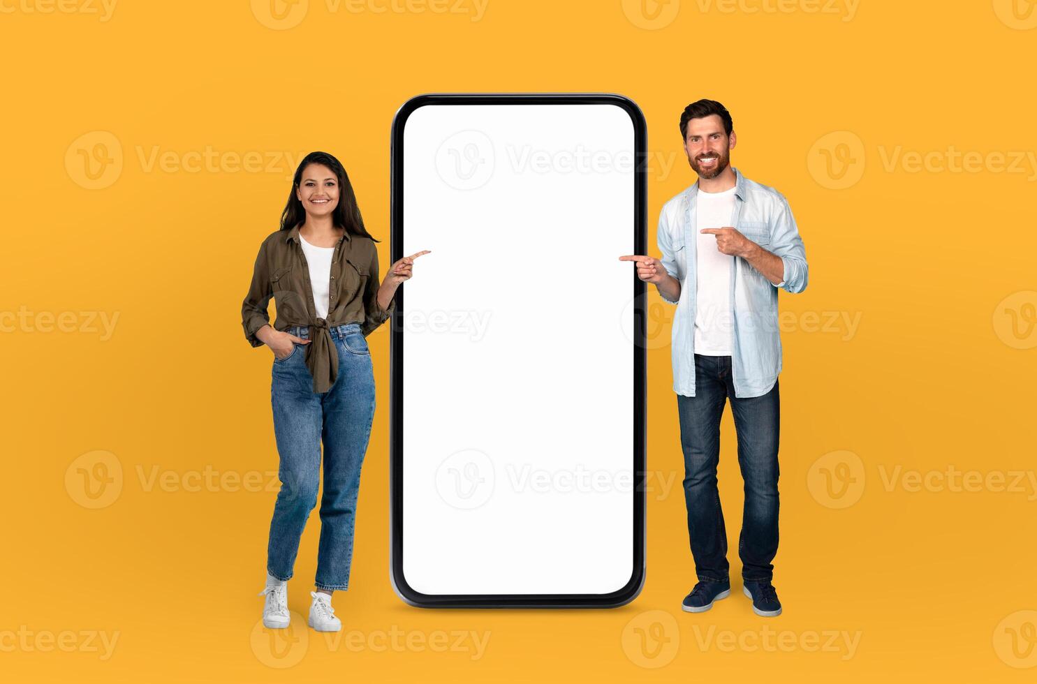 Multiethnic couple standing in front of a bright yellow background, both pointing at a blank screen. They seem focused and engaged photo