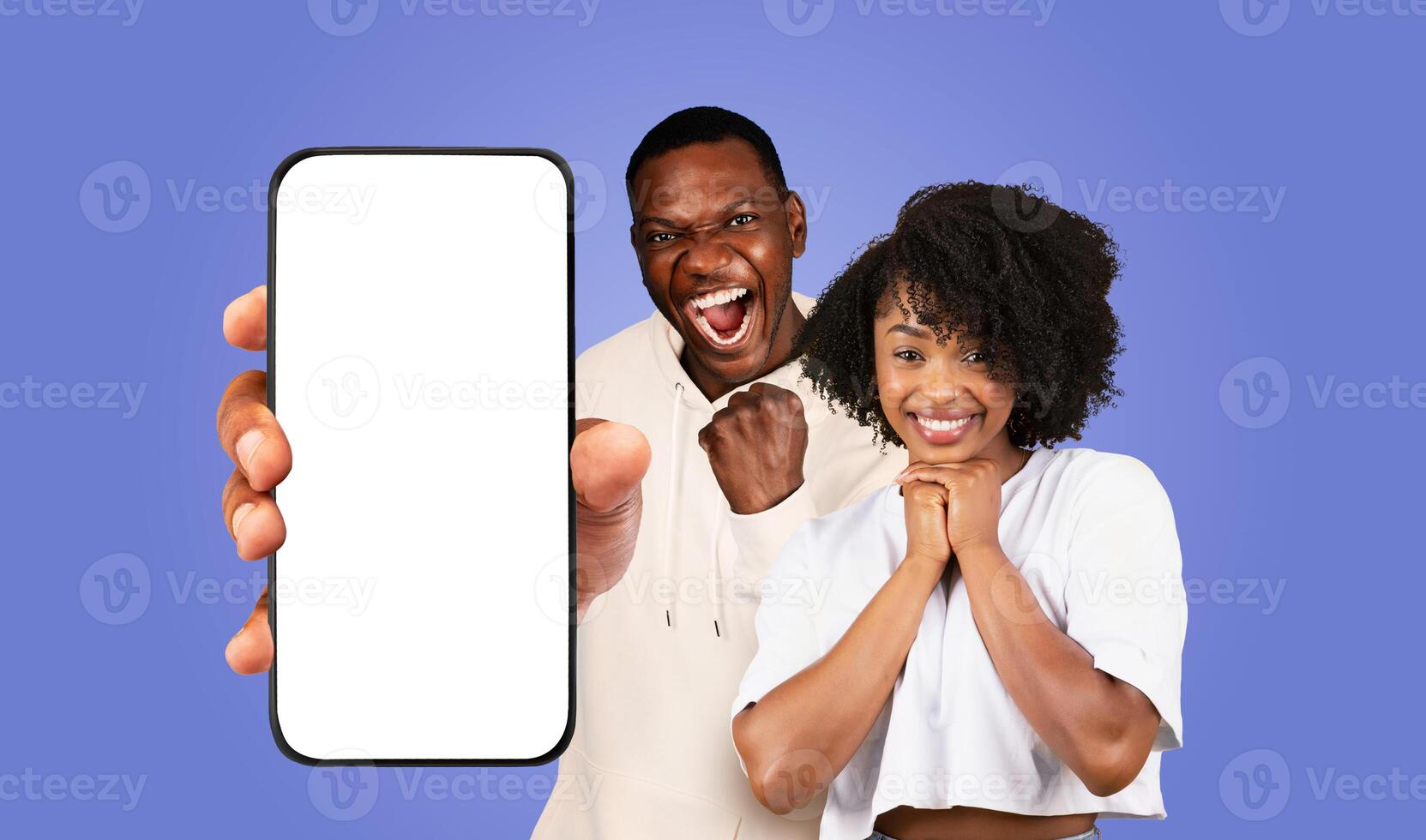 Black man and woman stand in front of a purple background. The man is holding a smartphone with a blank white screen, woman is smiling and has her hands clasped together in front of her. photo