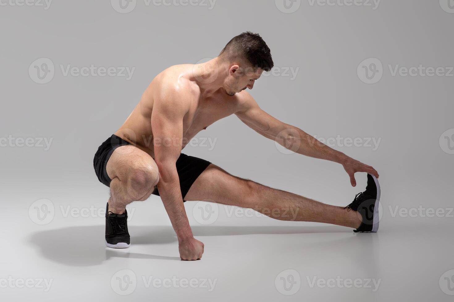 A fit, young man is seen stretching his leg muscles in a spacious, minimalist indoor gym, wearing black shorts and athletic shoes, practicing a deep lateral stretch to prepare for his workout routine photo