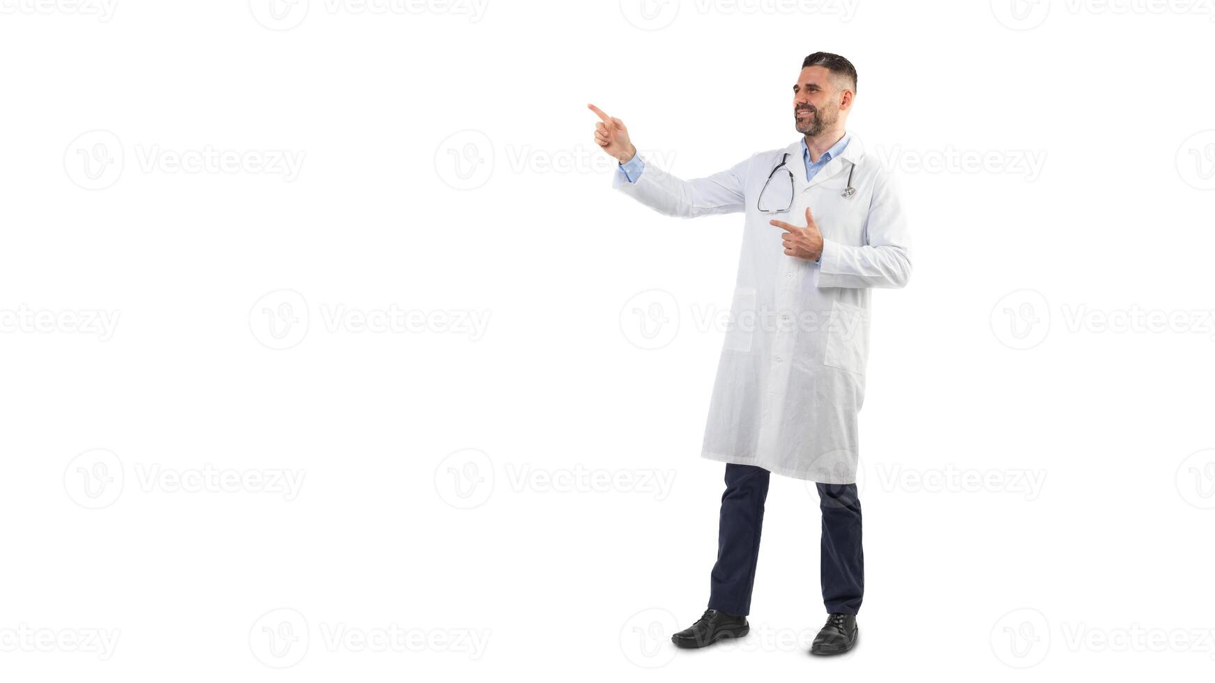 A man doctor is standing against a plain white background while pointing to the side and smiling. He is wearing a white lab coat with a stethoscope around his neck, copy space photo