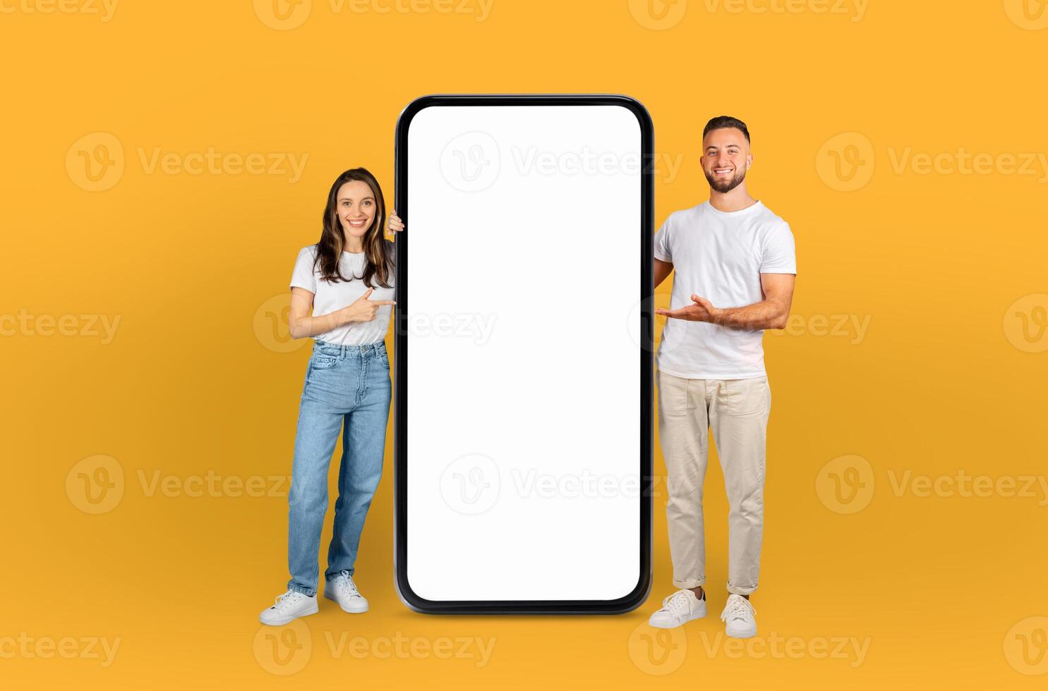 A man and woman are standing beside a blank phone screen, posing for a photo. They both appear to be smiling and looking at the screen, possibly preparing to take a selfie or share something photo