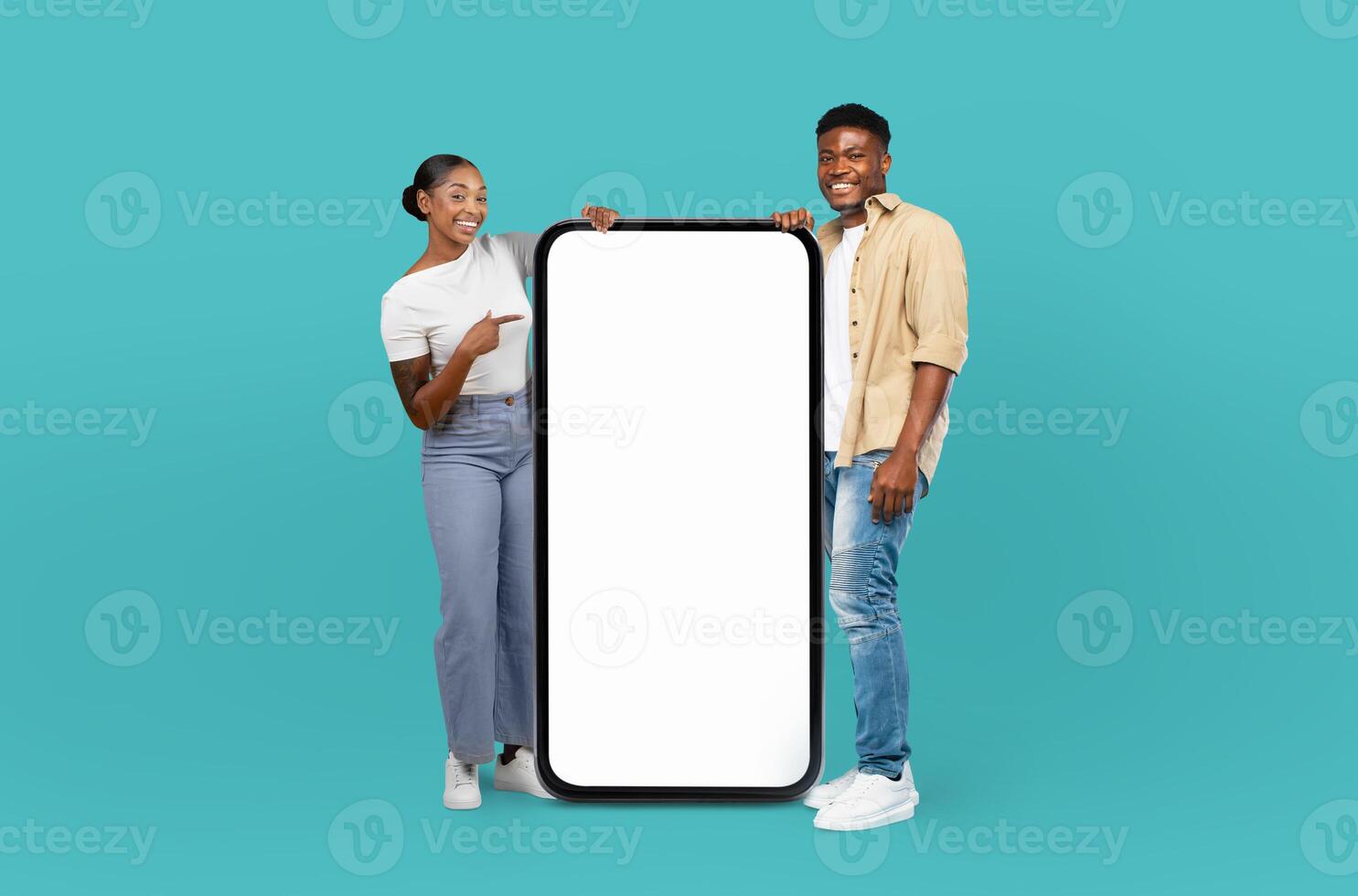 Black man and woman are standing against a blue background, holding up a blank screen. They appear to be showcasing something on the screen, potentially for a presentation or advertisement. photo