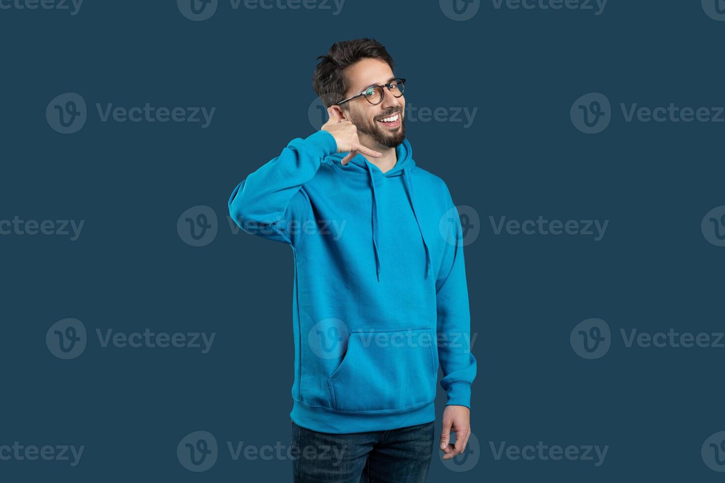 A cheerful guy wearing a bright blue hoodie stands confidently, flashing a warm smile. He makes a playful calling gesture with his hand near his ear photo