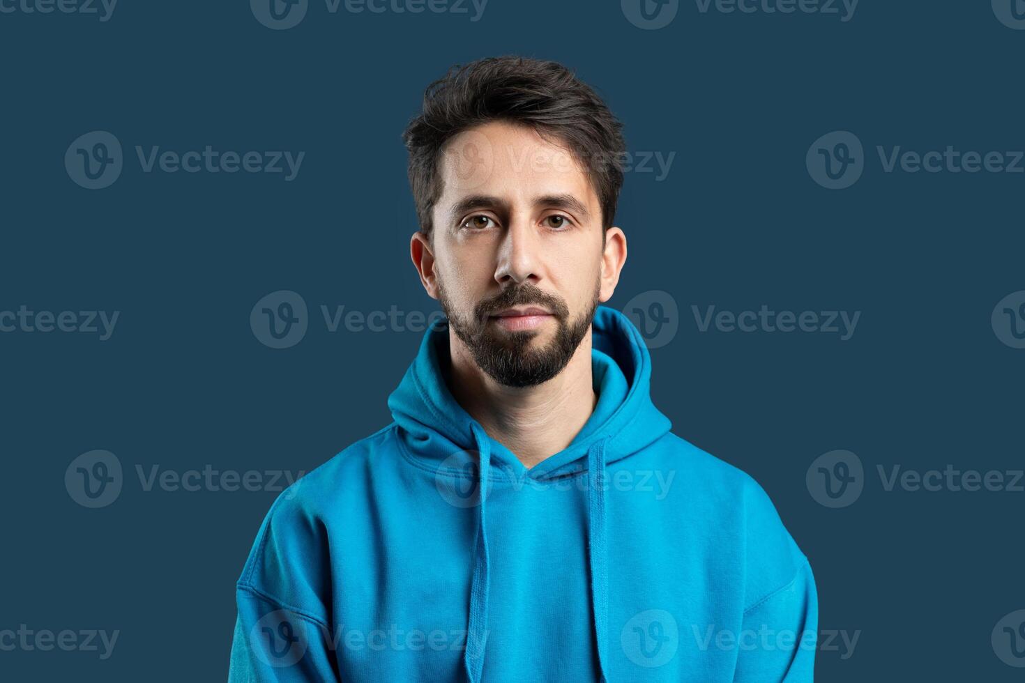 A man with a full beard is standing while wearing a blue hoodie. He appears relaxed and casual, with his hands in his pockets. The background is unremarkable, allowing the man to stand out. photo