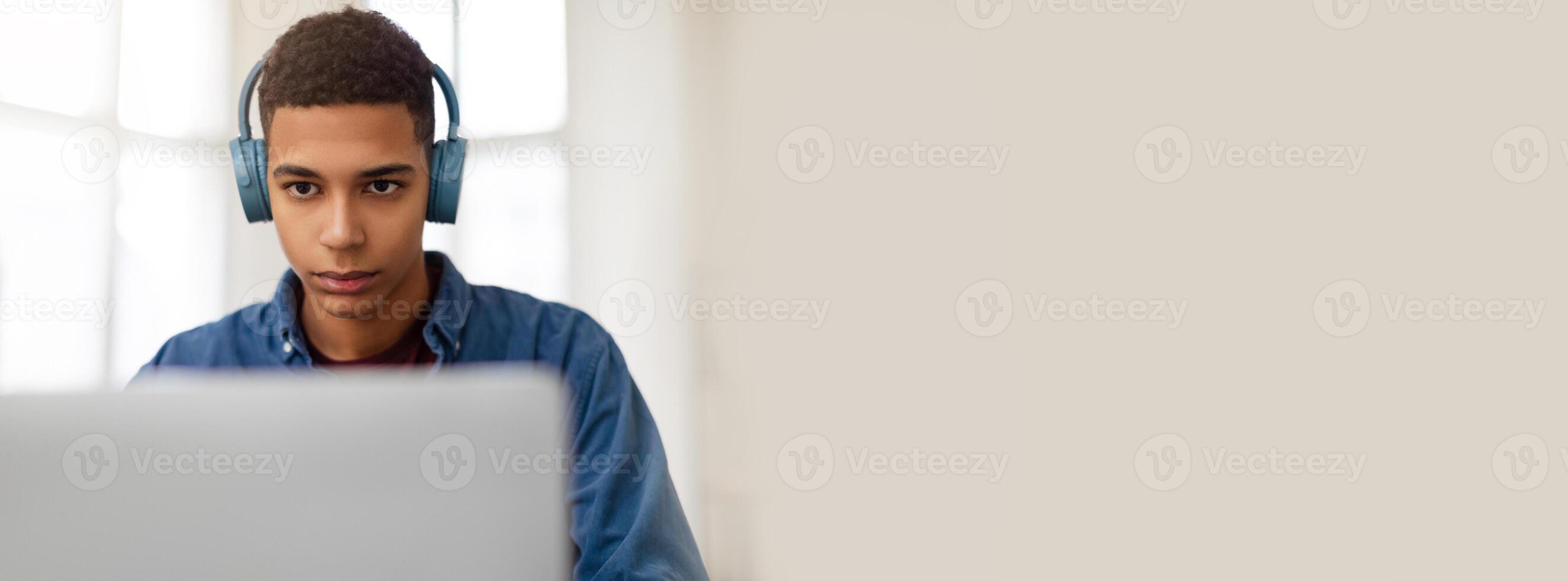 A young African American man is deeply engaged in his computer work, wearing headphones. The bright room features large windows that provide natural light, enhancing his concentration. photo