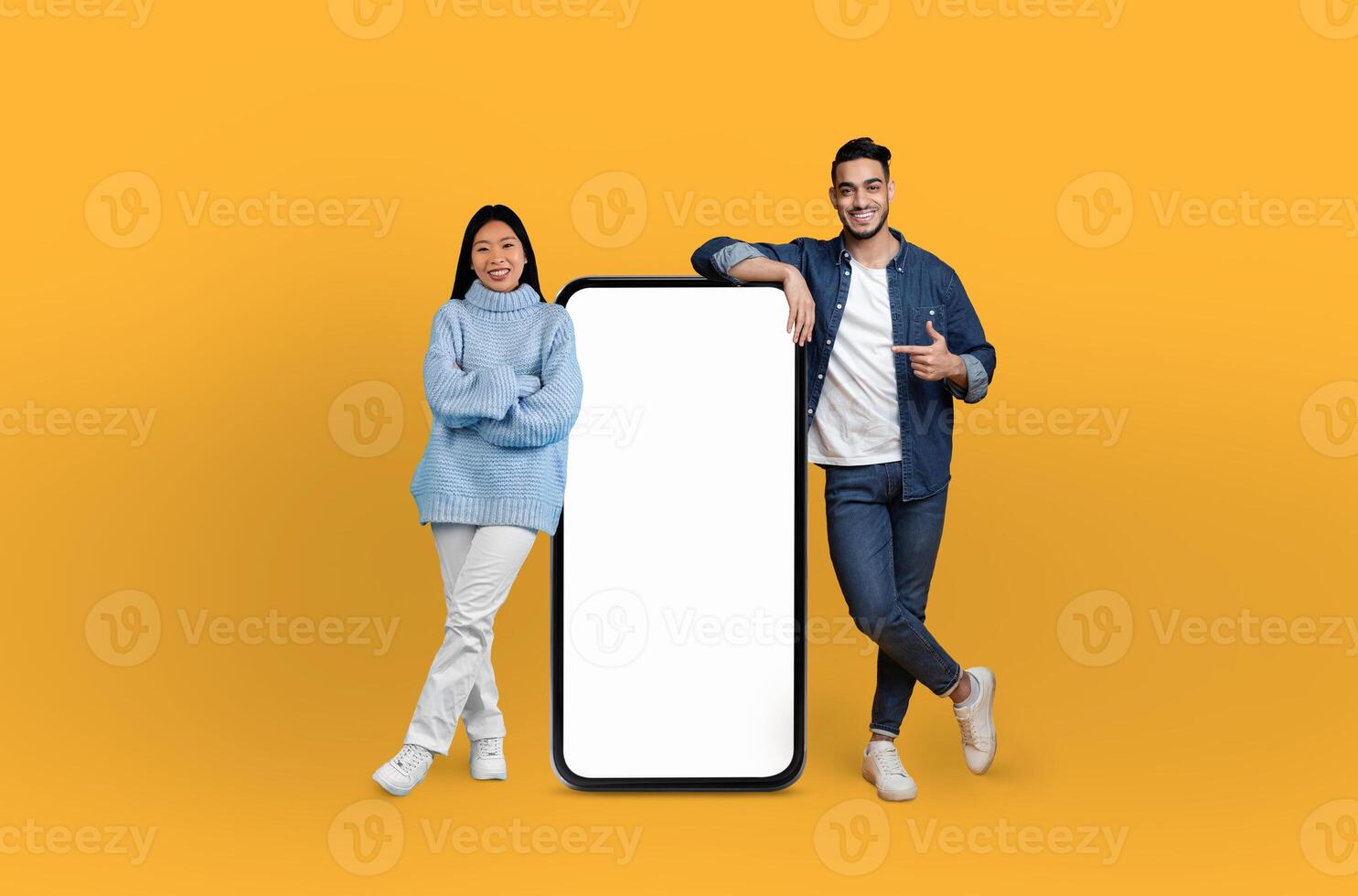 Multiracial man and woman standing in front of blank phone screen, striking pose and looking directly at camera. Phone screen is void of any content, emphasizing duos interaction with technology. photo