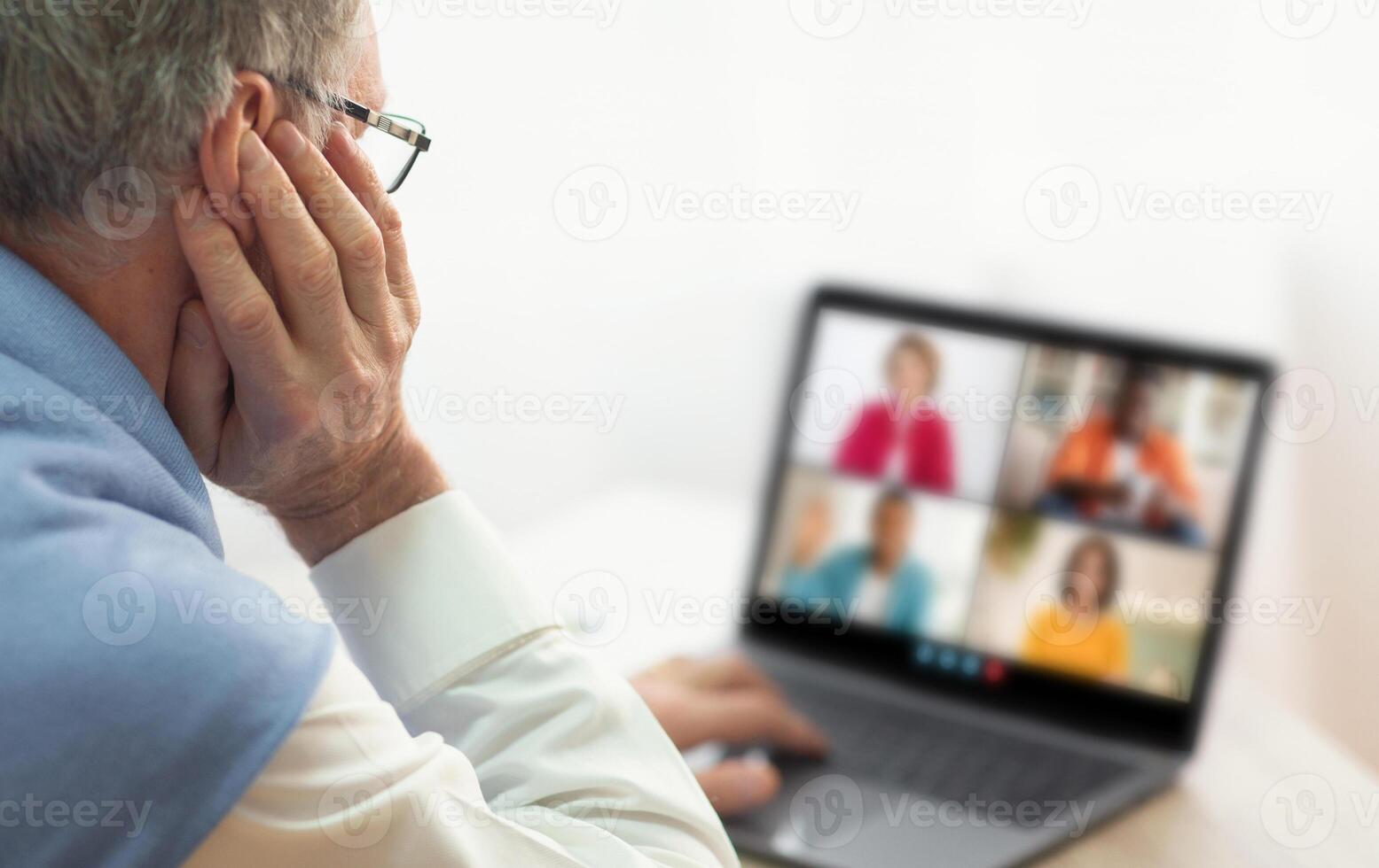 An older man with glasses is seen participating in a conference on a laptop. He is seated and his hand is placed to his ear, suggesting he is listening attentively photo