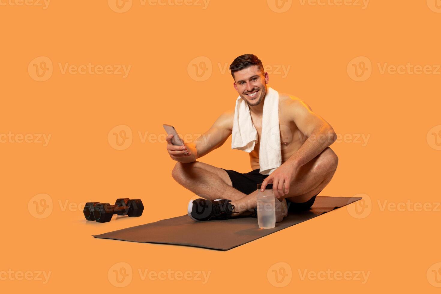 A man is seated on a mat, holding dumbbells in his hands while looking at his phone. The man appears to be engaged in a workout or exercise routine, possibly monitoring his progress photo
