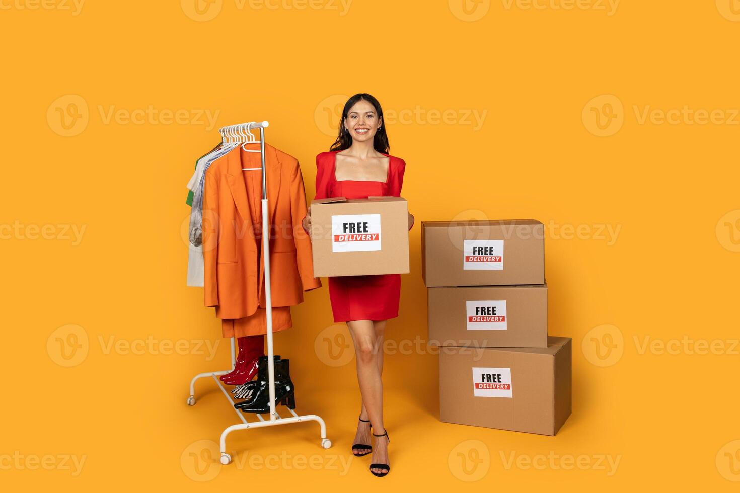 A woman dressed in a vibrant red dress stands confidently next to stacks of clothing boxes. She appears to be inspecting the items, possibly in a store or warehouse setting. photo