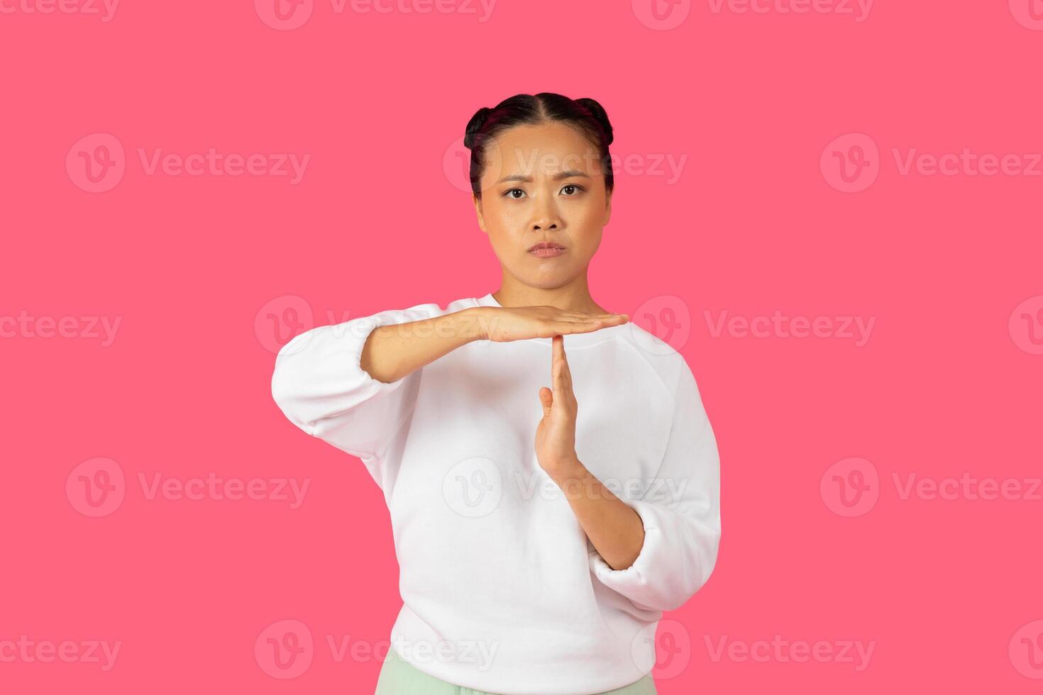 Asian woman is performing a stop gesture with her hands, expressing authority and confidence against a bright pink background. photo