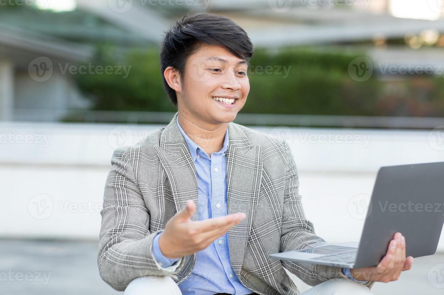 Asian young man is sitting outside on a bright day and smiling while using his laptop computer. He is wearing a plaid blazer, having a conversation, possibly over a call. photo