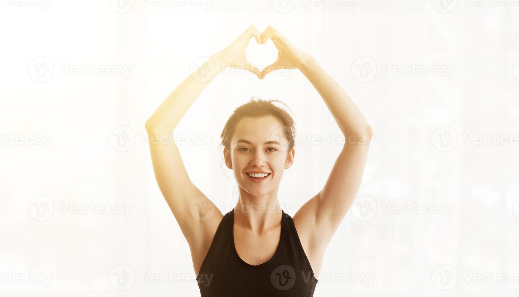 A woman standing with her hands raised above her head, forming the shape of a heart with her fingers. She appears to be smiling and expressing positivity and love through her hand gesture, sun flare photo