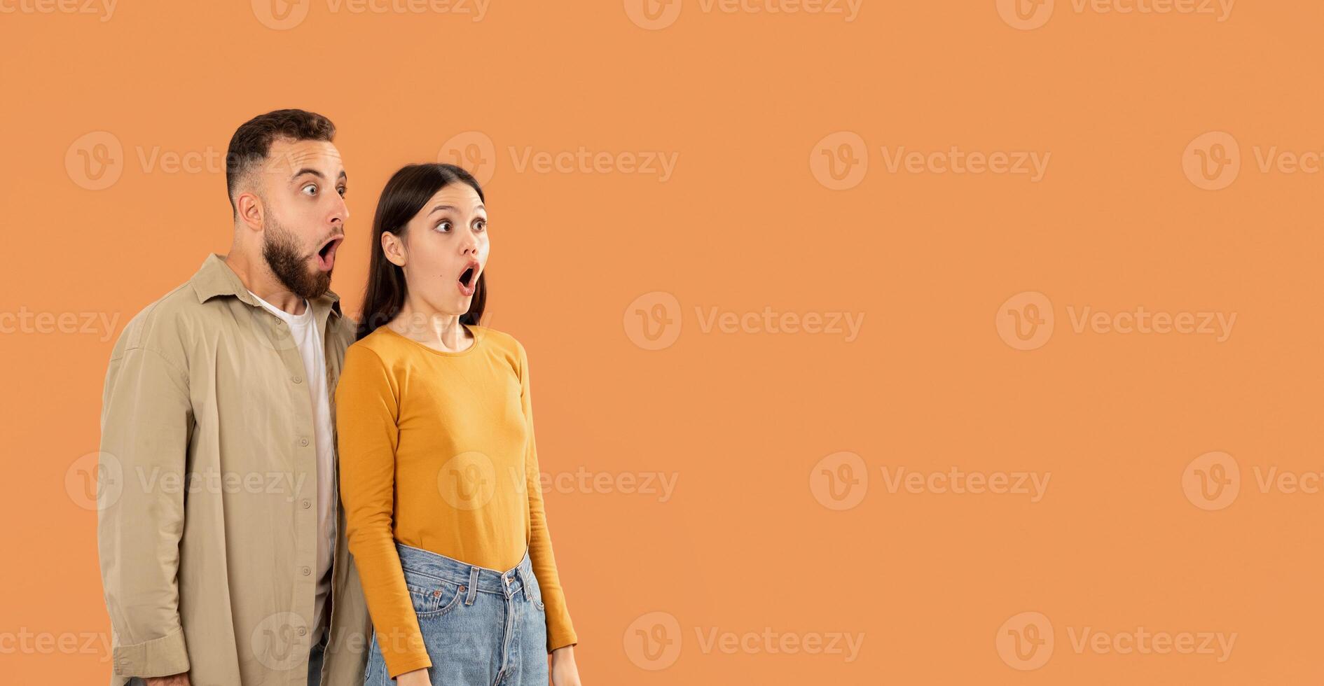 A young couple stands close together, displaying shocked expressions with wide-open mouths, against a solid orange background, panorama with copy space photo