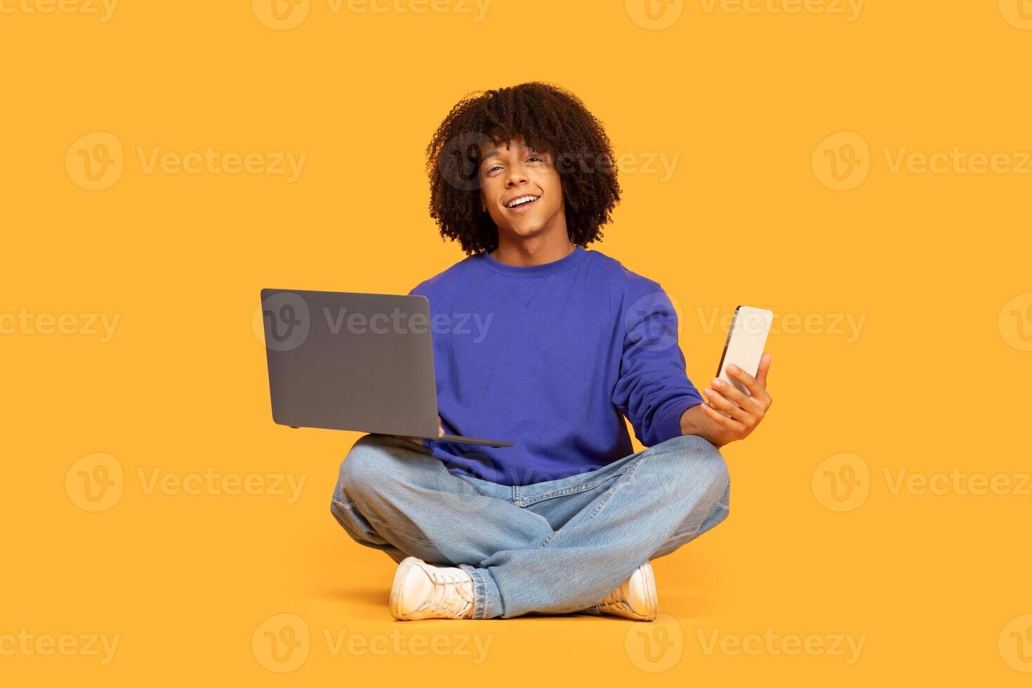 A young guy sits cross-legged on a vibrant yellow background, actively participating in online learning. He hold a smartphone in one hand and operate a laptop with the other photo