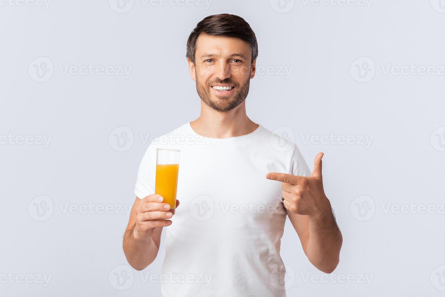 A man in a white t-shirt holds a glass of orange juice and points to his right, smiling at the camera. photo