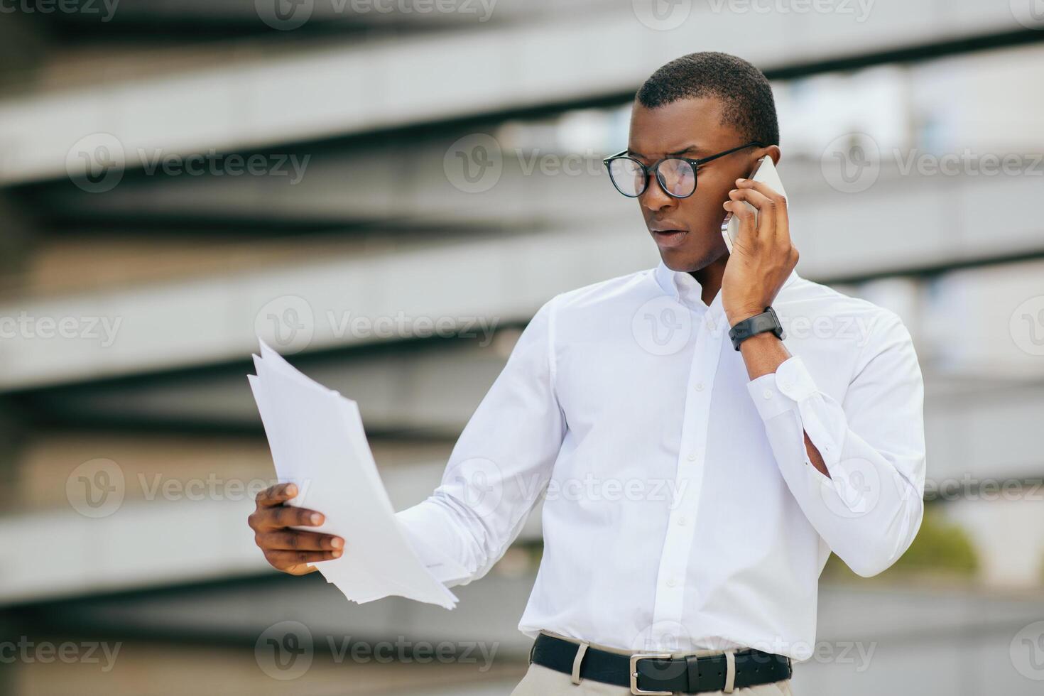 African American businessman in a white shirt and glasses is standing outside, holding papers in his left hand and talking on his phone with his right hand. He is looking down at the documents photo