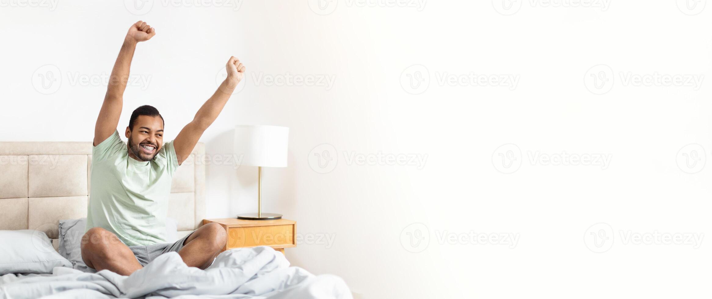African American man is sitting on the edge of his bed with arms raised, stretching as he greets the morning. The room is bright and cozy, creating a cheerful atmosphere. photo