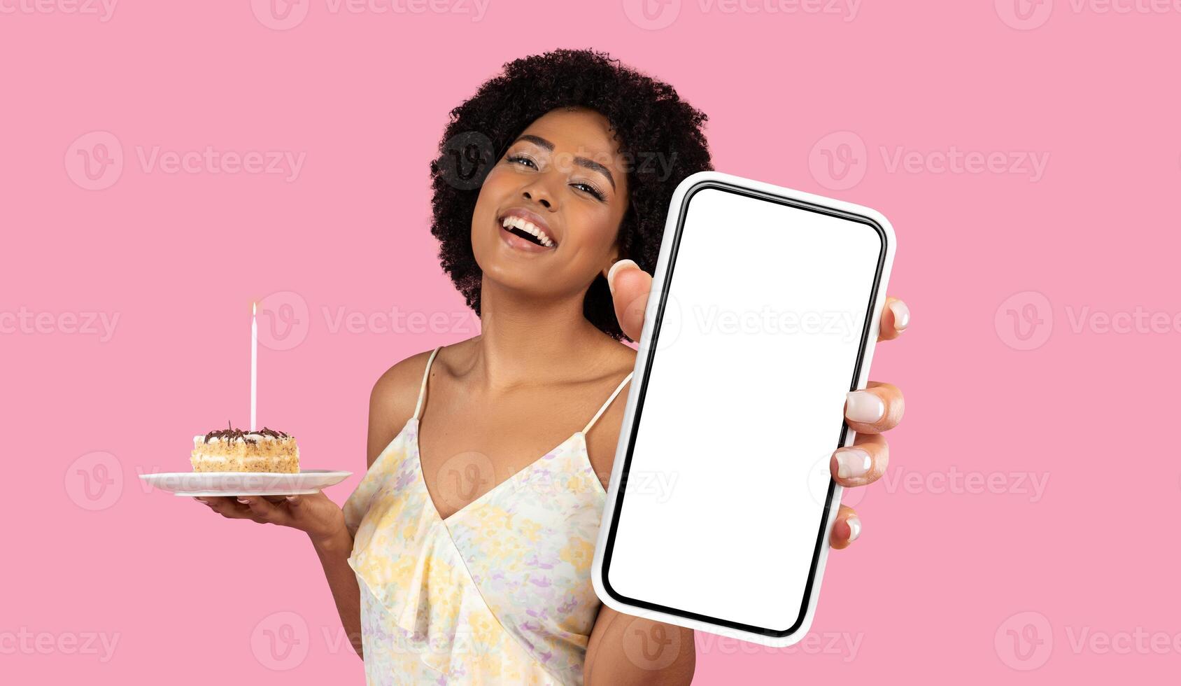 African American young woman with curly hair is holding a piece of birthday cake in one hand and a smartphone with a blank screen in the other hand. The background is a solid pink color. photo