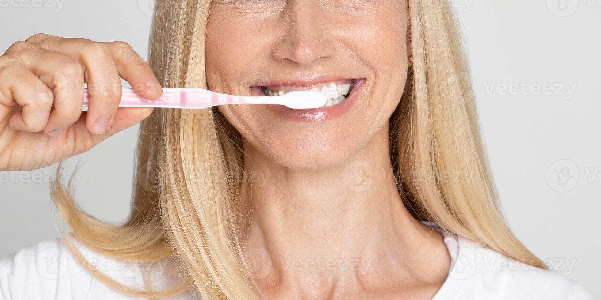 A close-up image of a woman with blonde hair smiling as she brushes her teeth with a pink toothbrush. The woman appears to be in her late 40s or early 50s, and her white teeth are visible photo