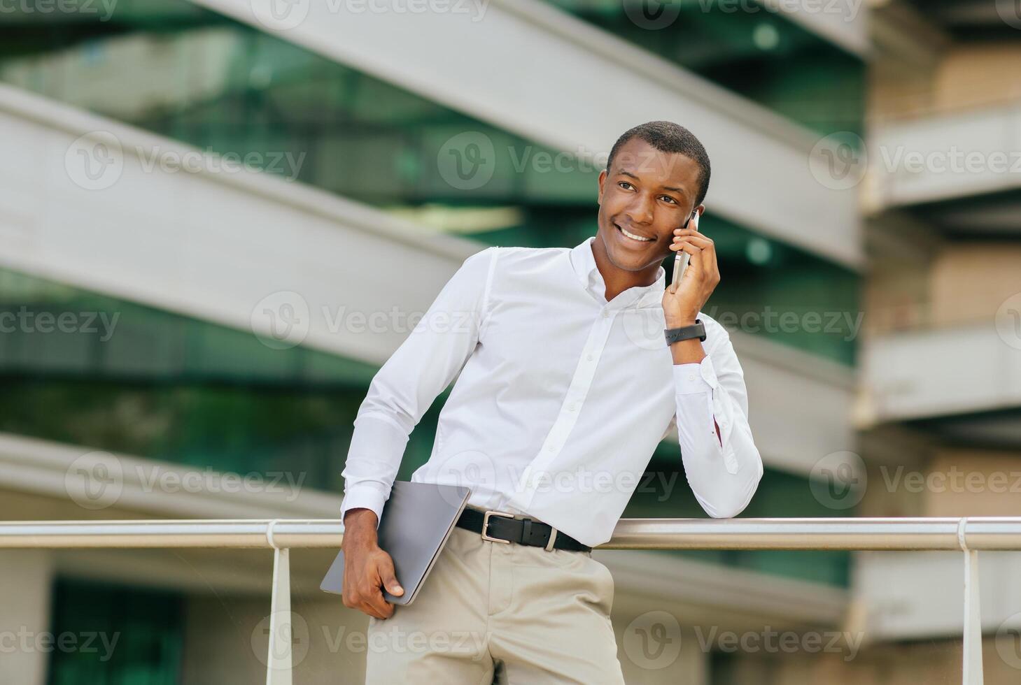 Black businessman, dressed in a white shirt and khaki pants, stands outside a modern office building and talks on his cell phone. He is holding a laptop under his arm and smiling photo