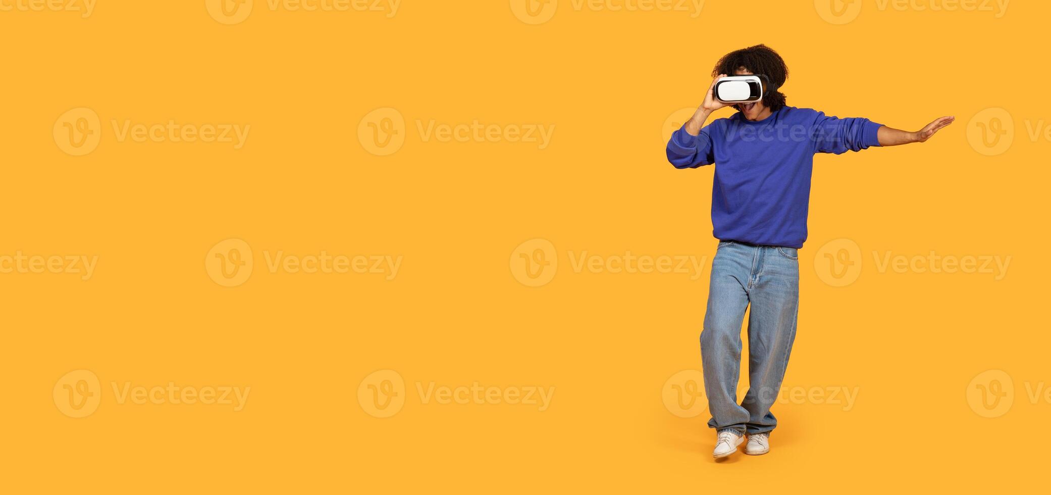 A young man stands against a vibrant yellow backdrop, fully immersed in a virtual reality experience while wearing a VR headset, copy space photo