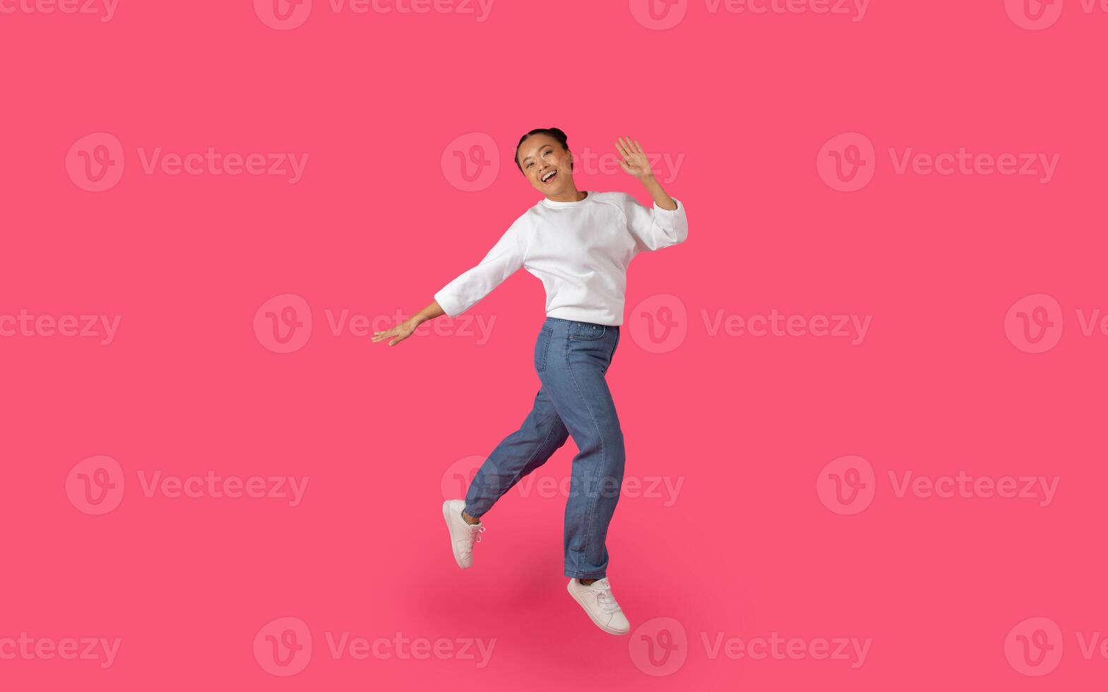 Asian young woman jumps and smiles in front of a bright pink background, her arms outstretched and her white shirt billowing in the air. photo